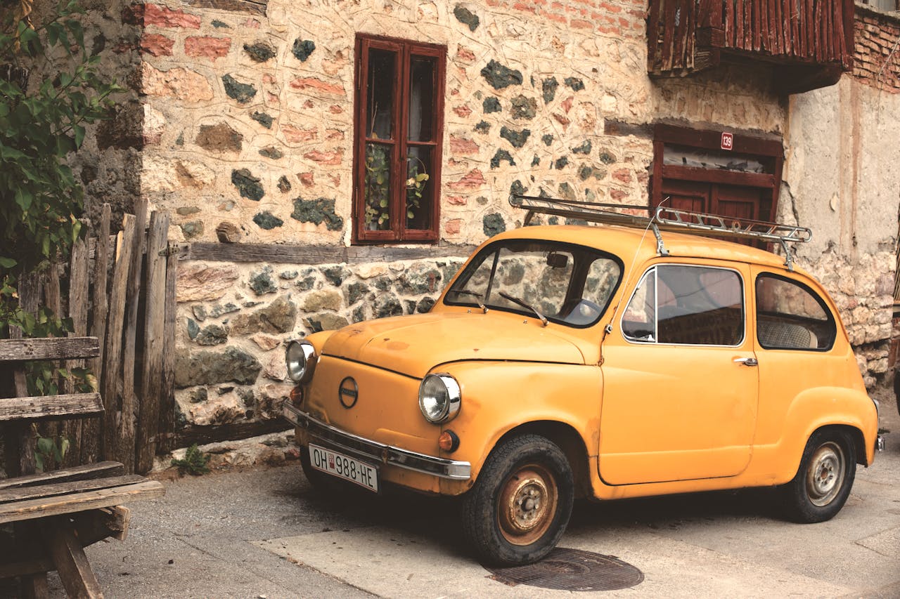 Old yellow car parked by a rustic stone building embracing vintage charm.